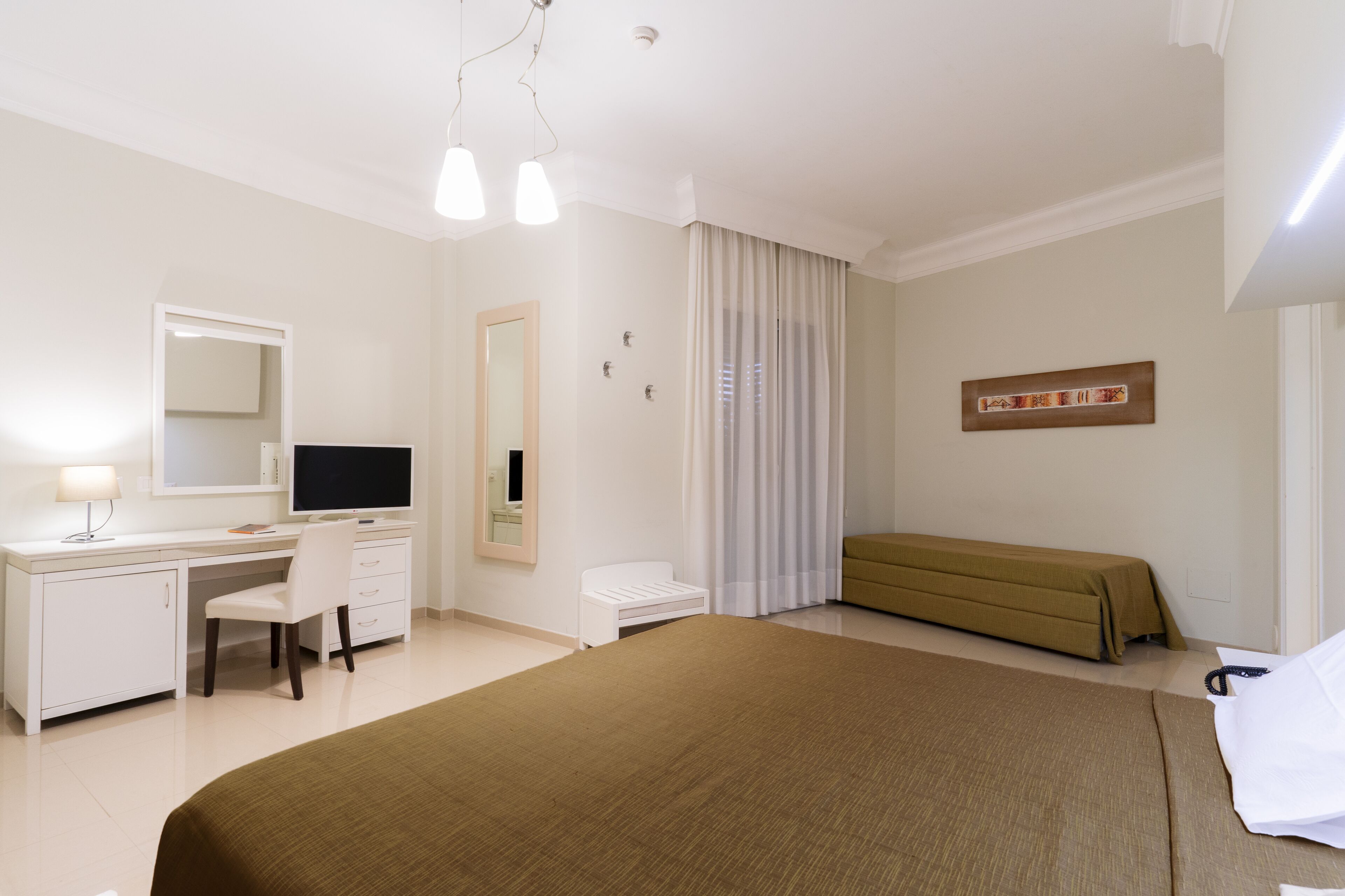 A hotel room with a bed, a desk with a computer, a chair, a bedside table, and a window with curtains.