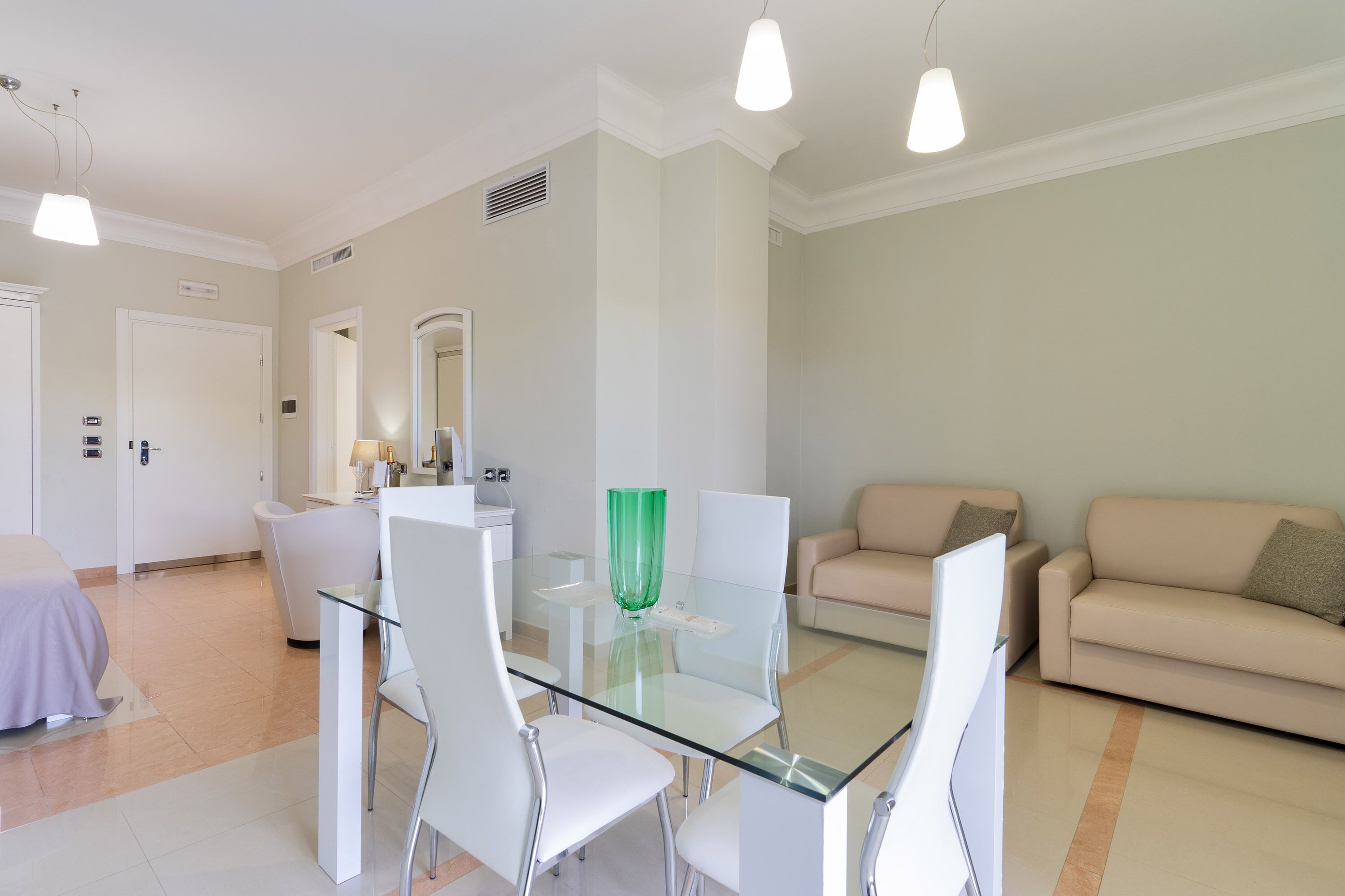 Un salon moderne avec une table à manger en verre, des chaises blanches, un canapé beige et un grand miroir.