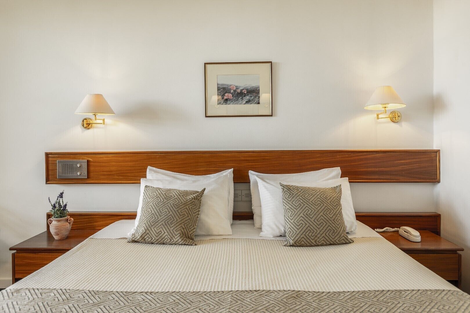 Una cama bien hecha con dos almohadas, una lámpara de mesita de noche, un teléfono y una pequeña planta sobre una mesita de madera.