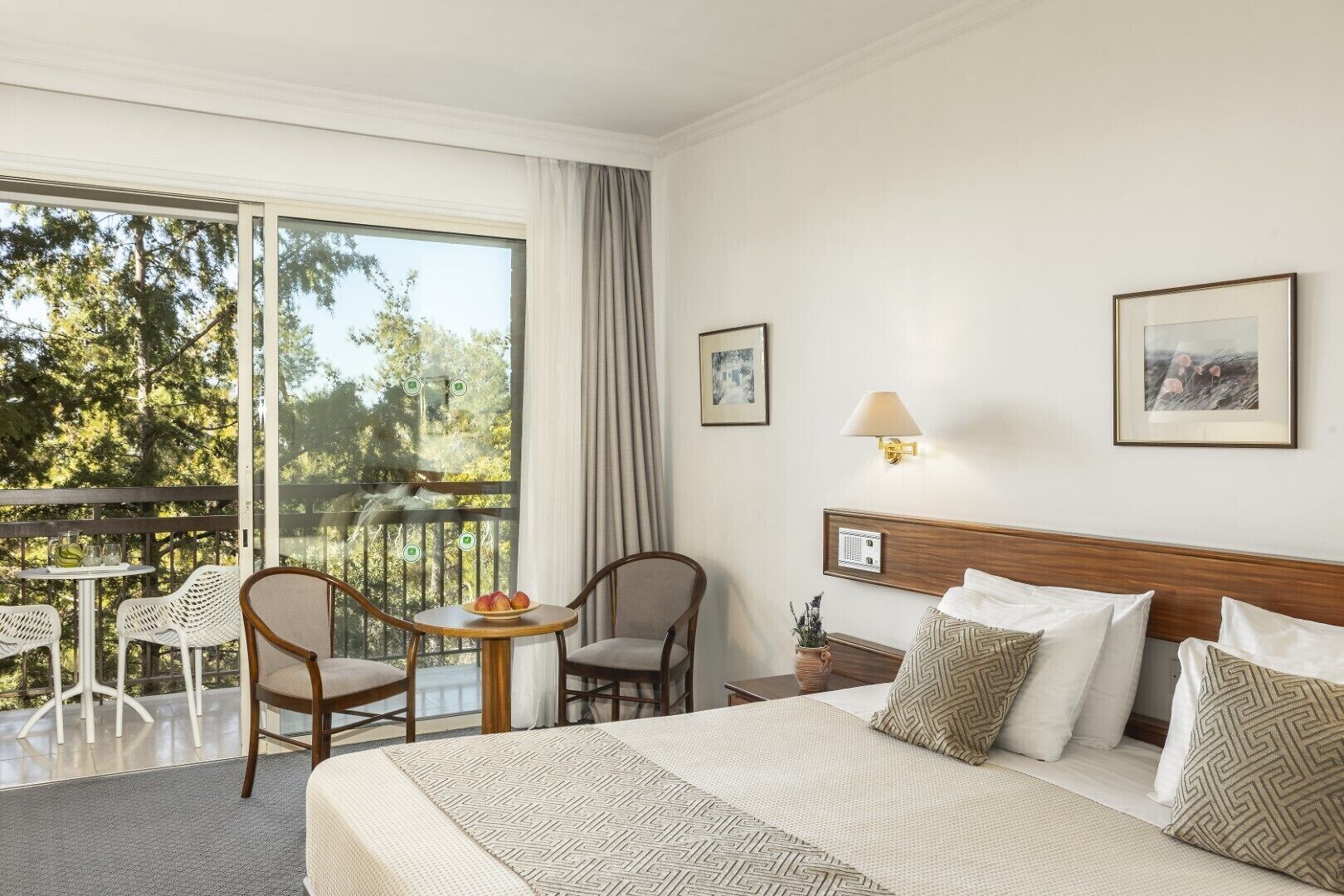 Habitación de hotel con cama, dos sillas, una mesita con fruta, balcón con mesa y sillas, y dos cuadros en la pared.