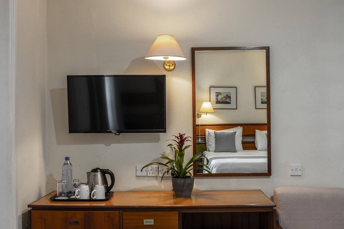 A hotel room with a flat-screen TV, a bedside lamp, a mirror reflecting the bed and a painting, a wooden desk with a kettle, cups, and a water bottle.