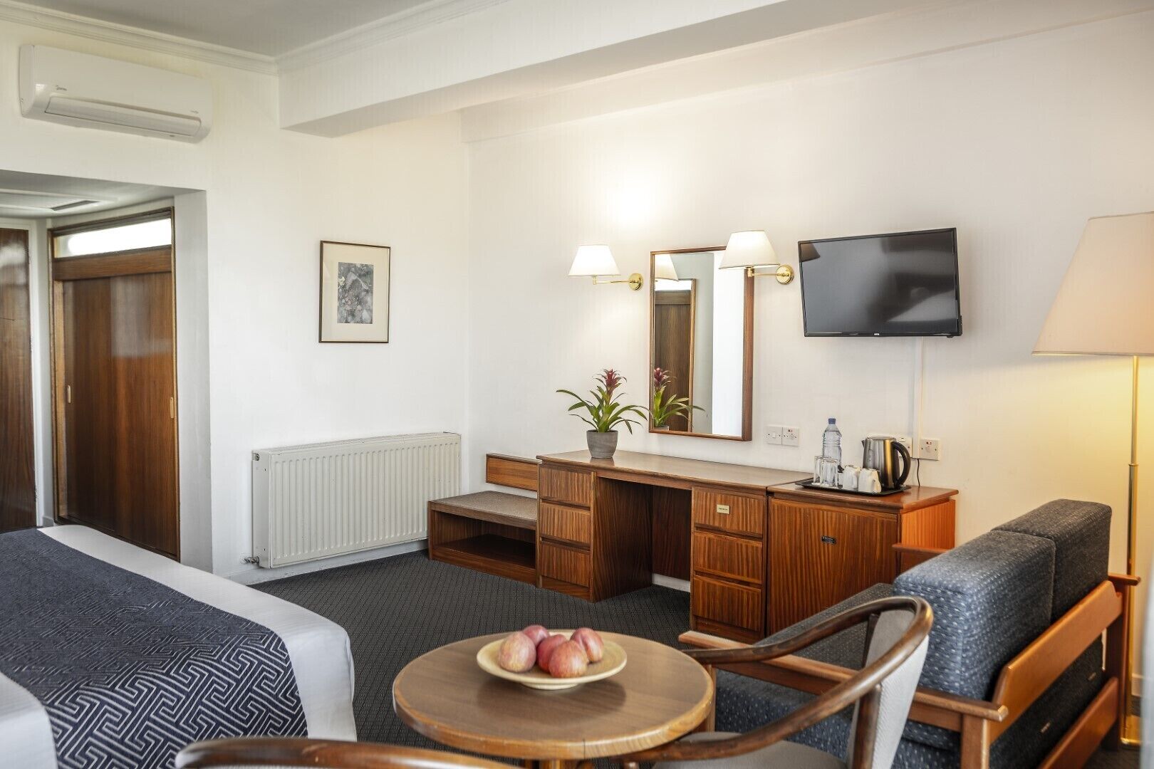 A hotel room with a bed, a desk with a mirror, a television, and a small table with fruit.