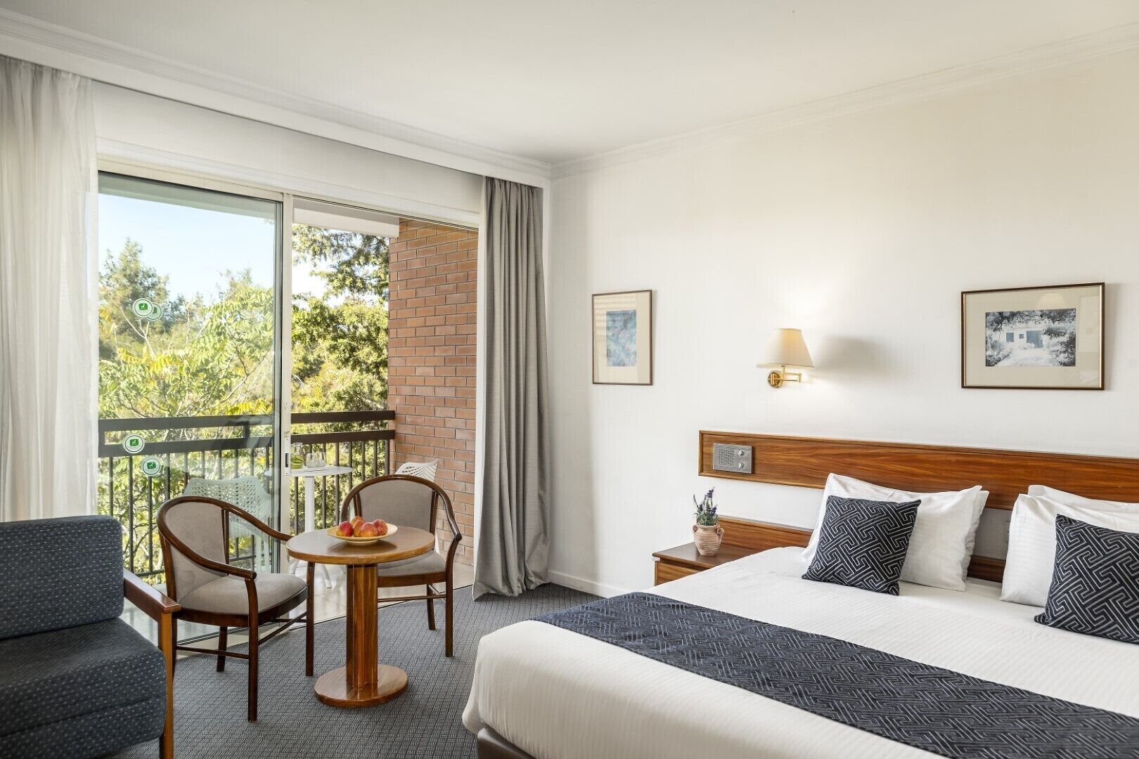 Habitación de hotel con cama, dos sillas, una mesita con fruta, un balcón con vistas a árboles y dos cuadros en la pared.