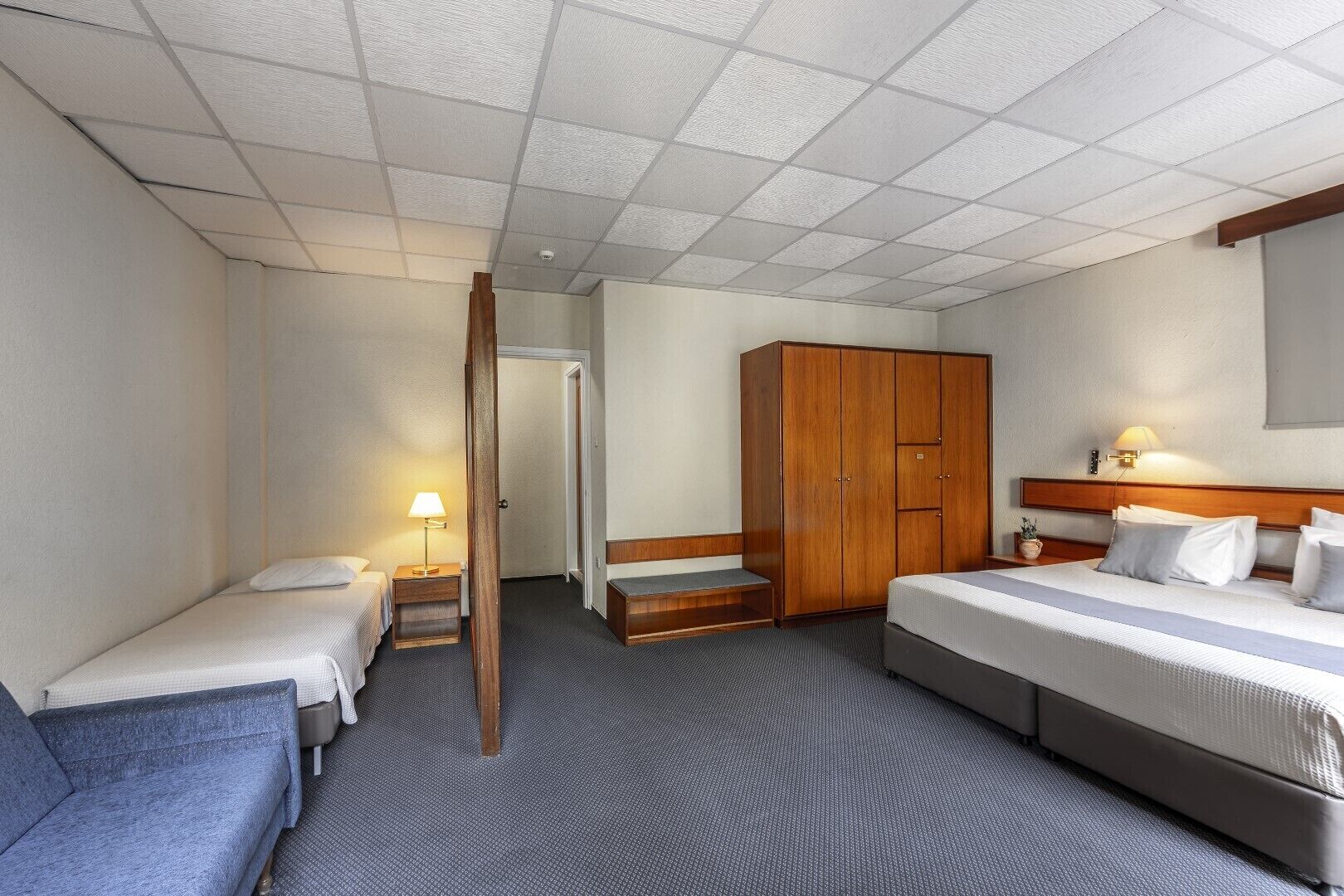 Quarto de hotel com duas camas de solteiro, uma cama de casal, um guarda-roupa, uma secretária com uma luminária e uma pequena mesa com uma planta.