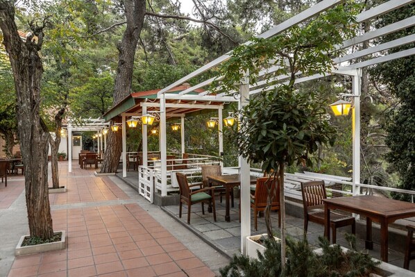 Un patio exterior con mesas y sillas de madera, una pérgola con luces colgantes y un sendero de ladrillo.