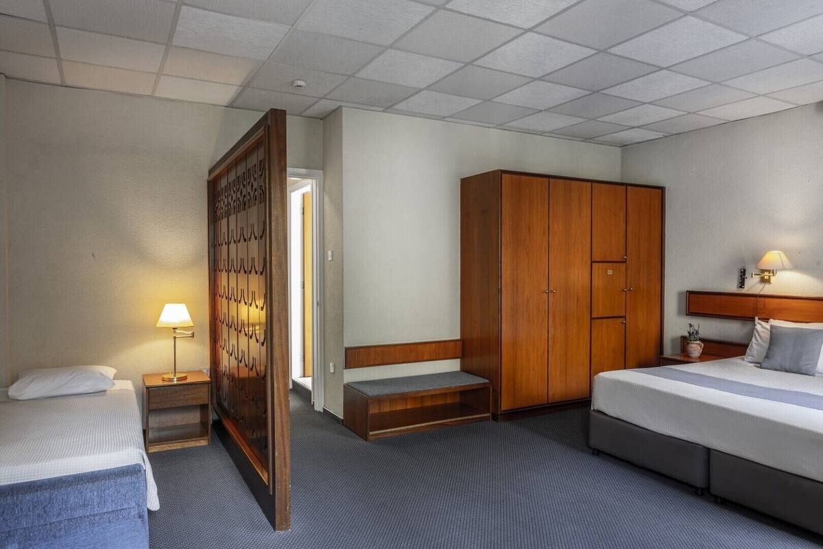 A hotel room with two beds, a wooden wardrobe, a bench, a nightstand with a lamp, and a wall-mounted light fixture.