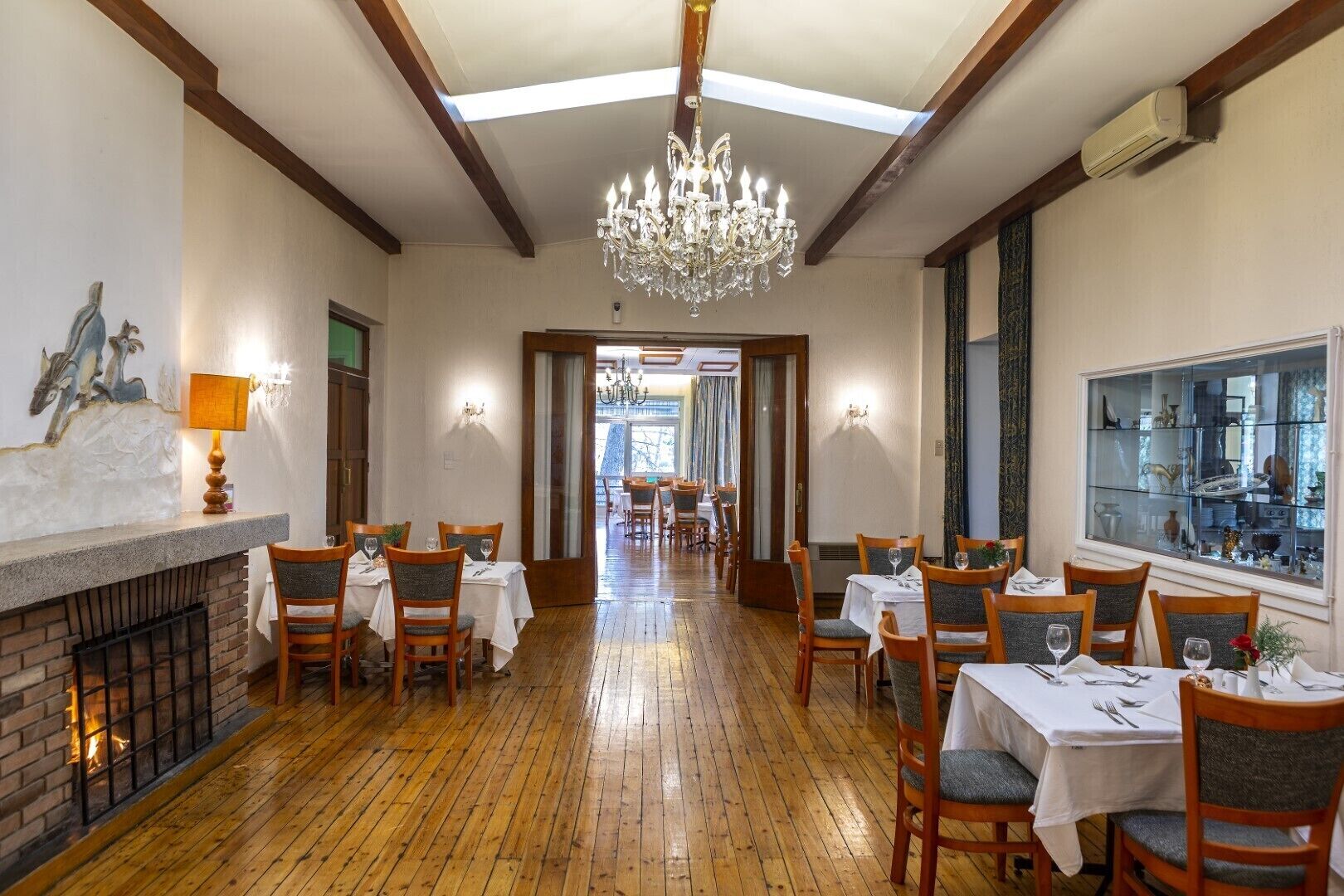 A dining area with wooden flooring, tables set with white tablecloths, and chairs. A chandelier hangs from the ceiling, and a fireplace is visible on the left.