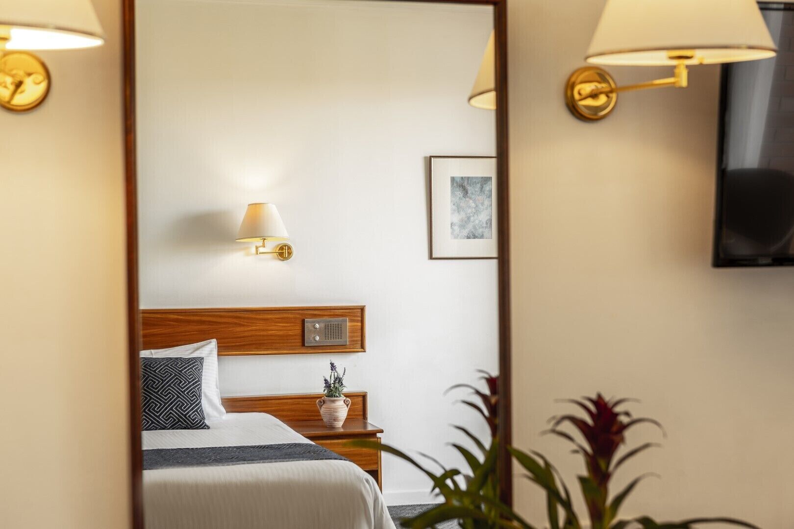 A hotel room with a wooden headboard, a bedside table with a vase, a framed picture on the wall, and a TV mounted above the bed.