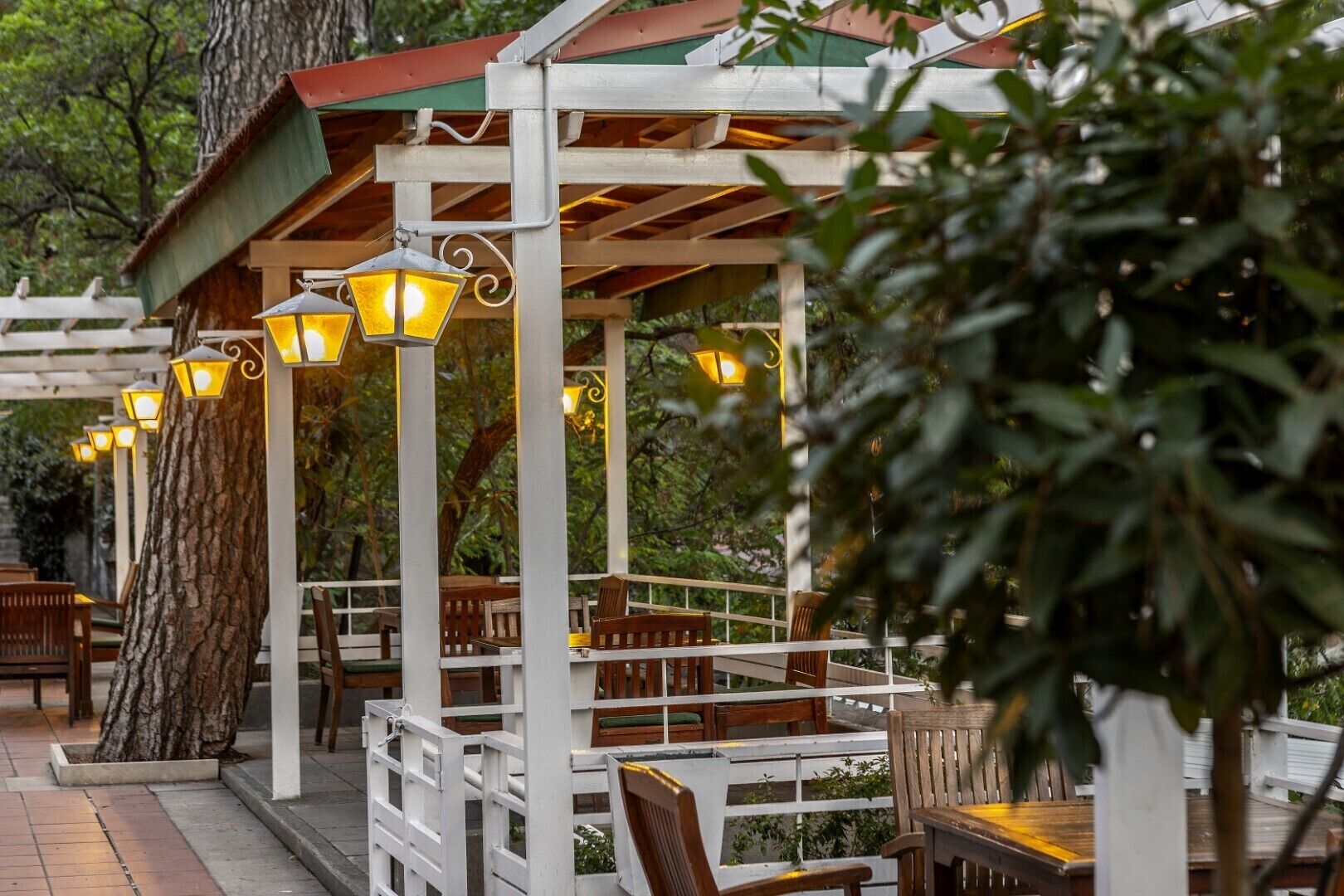 An outdoor seating area with wooden benches and tables, surrounded by white railings and illuminated by hanging lanterns.