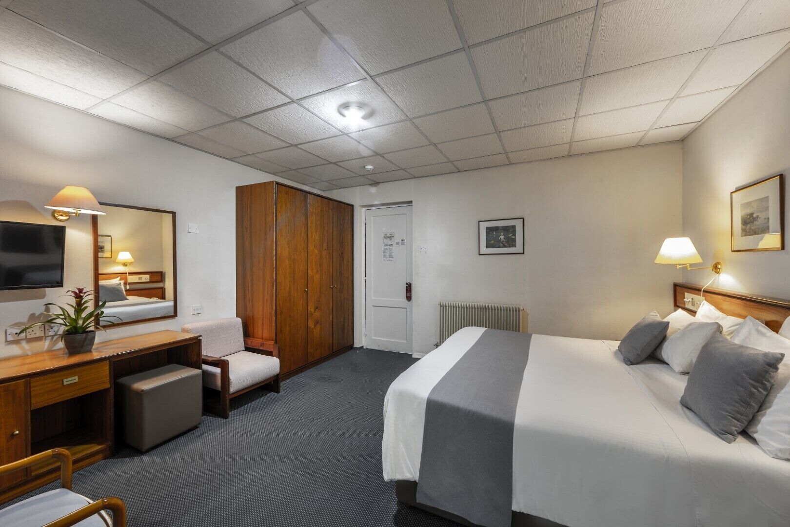 A hotel room with a bed, a desk with a chair, a wardrobe, a radiator, a television, and a framed picture on the wall.