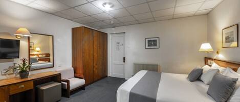 A hotel room with a bed, a desk with a chair, a wardrobe, a radiator, a television, and a framed picture on the wall.