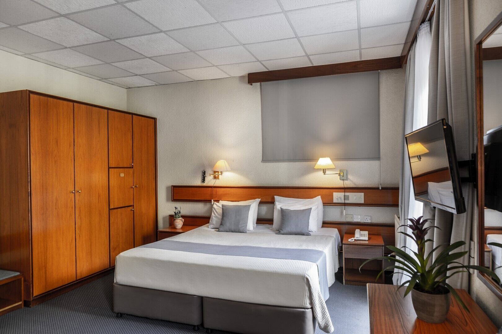 A hotel room with a large bed, wooden wardrobe, bedside lamps, a TV, and a potted plant.
