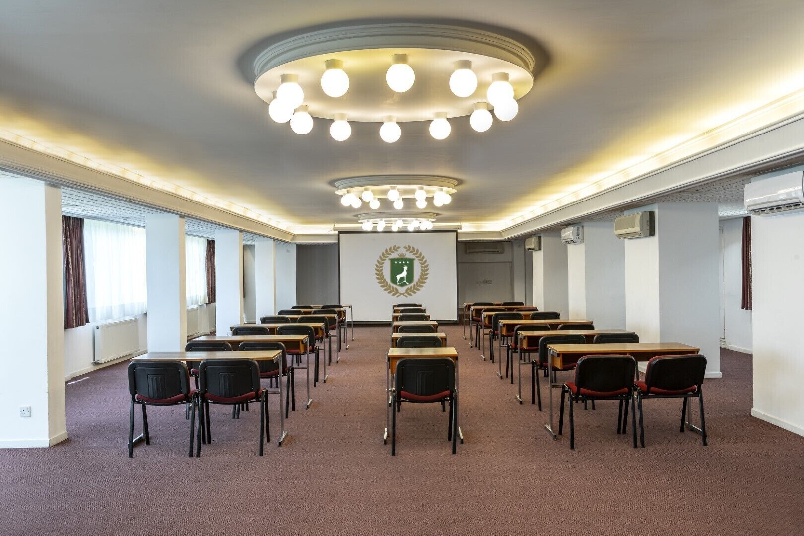 Ett konferensrum med rader av skrivbord och stolar, en projektorduk och en logotyp på väggen.