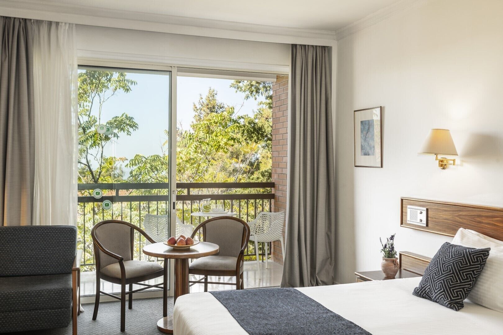 Habitación de hotel con cama, dos sillas, una mesita con fruta, un balcón con vista a árboles y una lámpara en la pared.