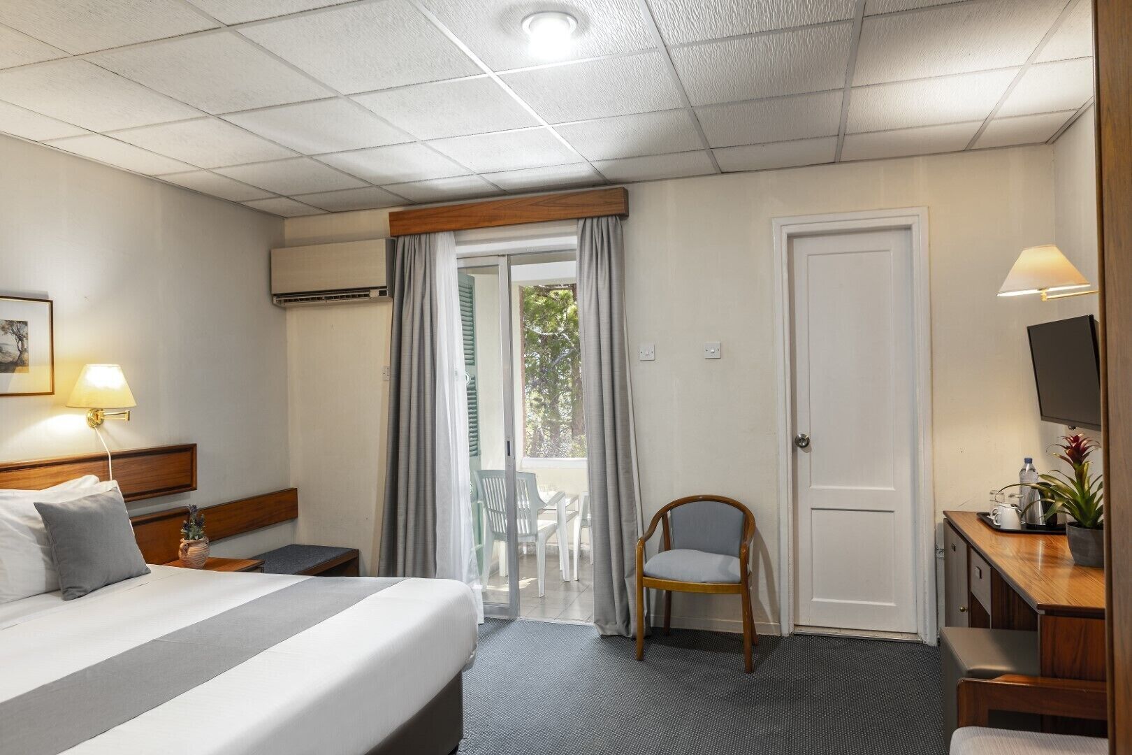 A hotel room with a bed, bedside table, lamp, chair, desk, TV, and a door leading to a balcony.