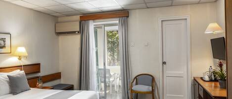 A hotel room with a bed, bedside table, lamp, chair, desk, TV, and a door leading to a balcony.