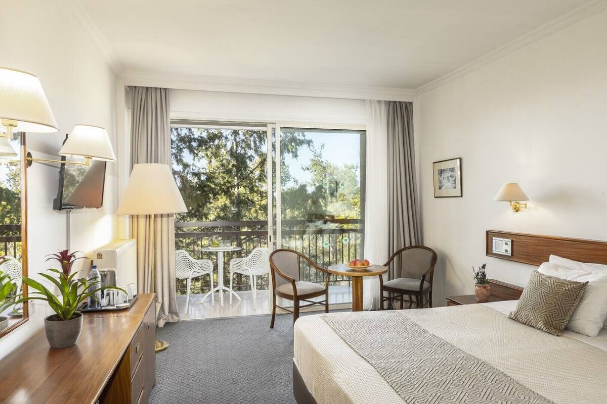 A hotel room with a bed, a desk with a lamp, two chairs, a small table with fruit, and a balcony with a view of trees.