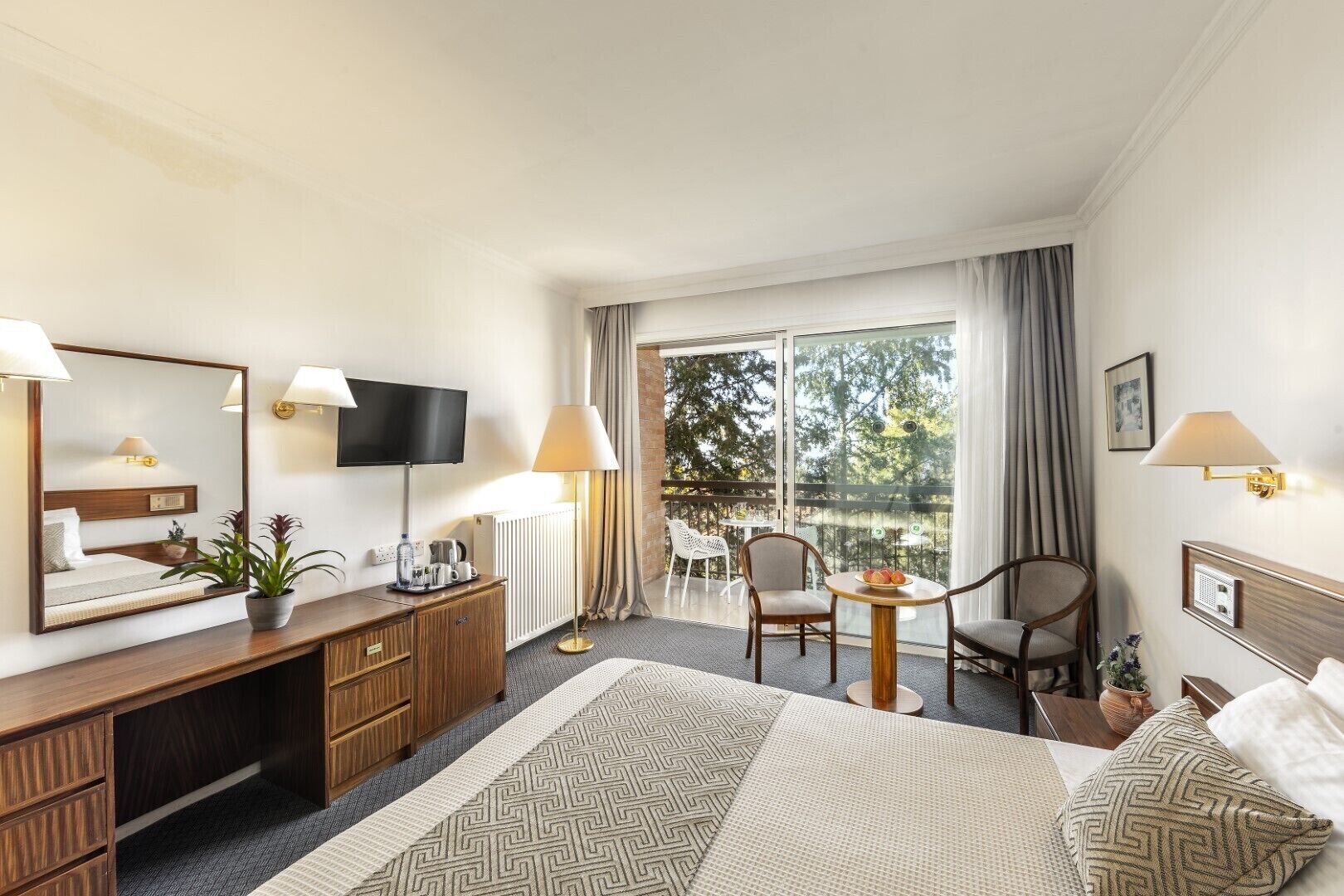 Habitación de hotel con una cama grande, un escritorio con espejo, televisión, balcón con vistas a árboles y un comedor con sillas.