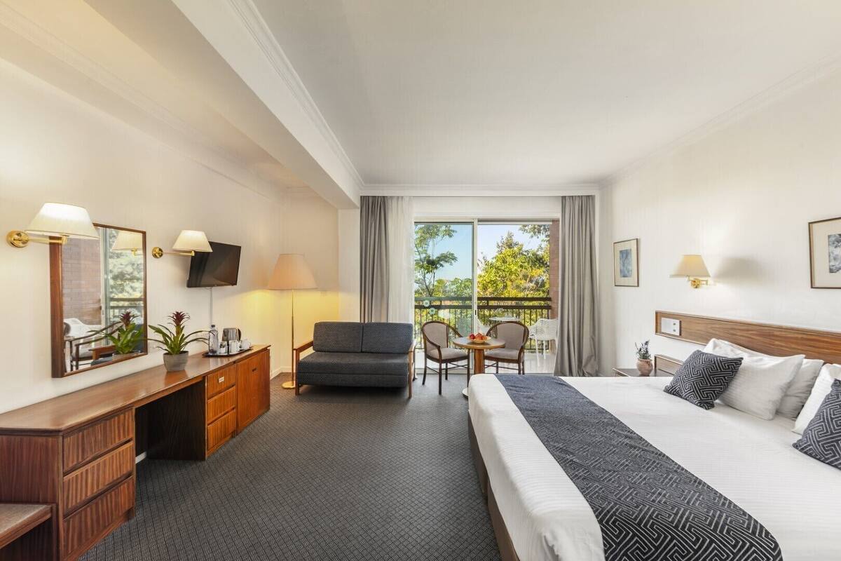 A hotel room with a bed, a desk, a chair, a TV, a balcony with a view of trees, and a window with curtains.