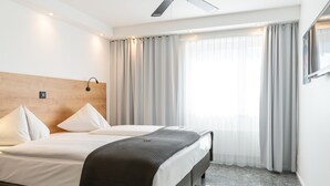 A hotel room with a bed, two pillows, a bedside table, a lamp, a ceiling fan, and a window with curtains.