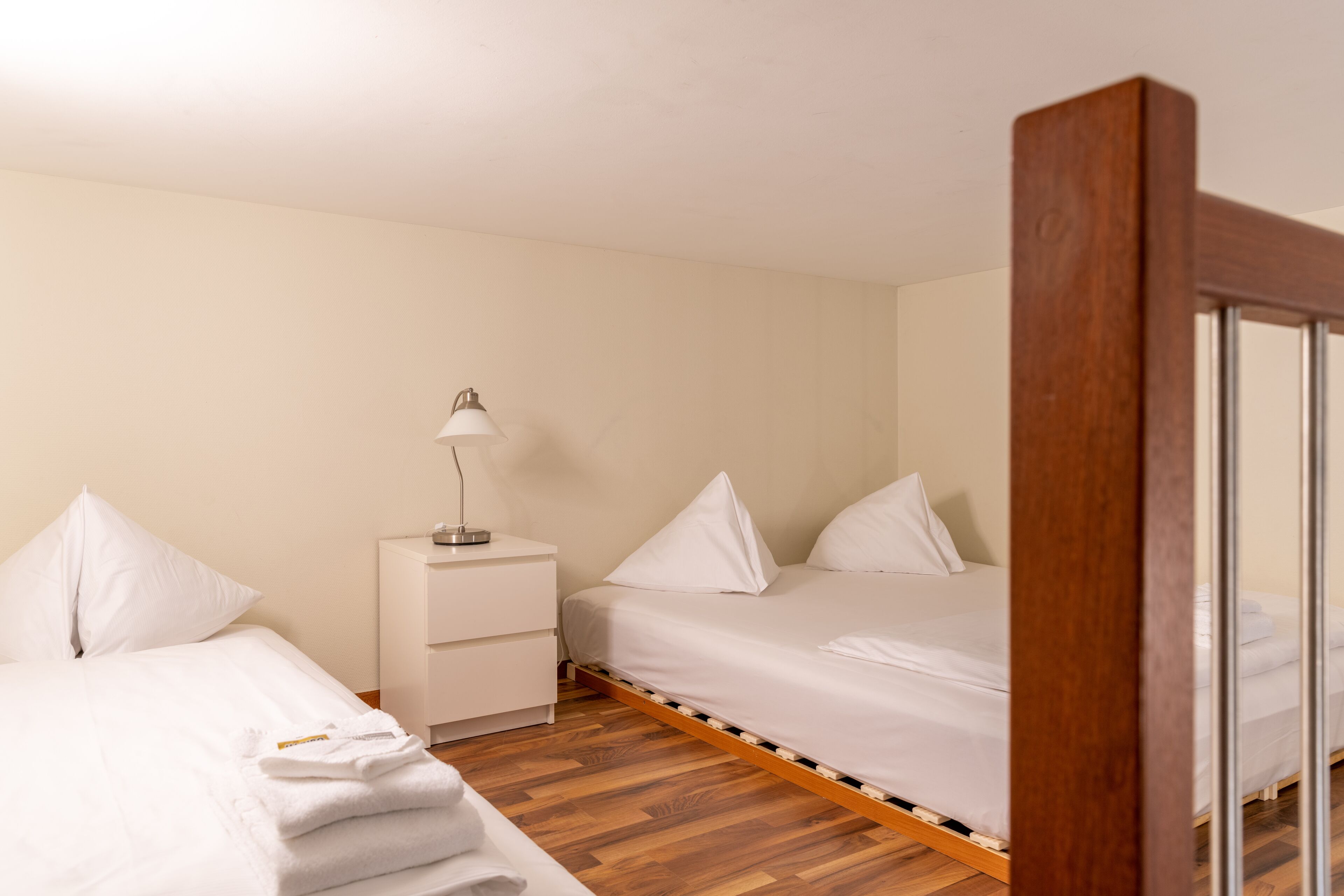 A hotel room with two beds, a bedside table with a lamp, and a wooden floor.