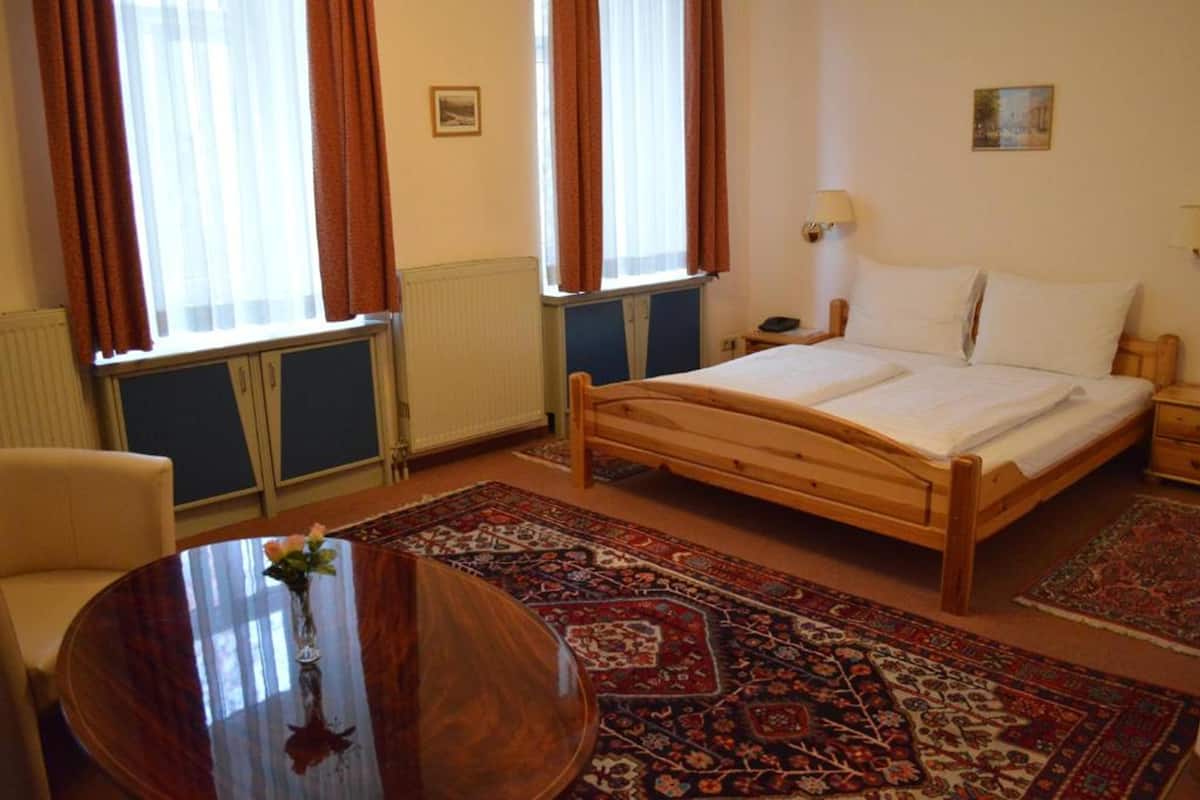 A hotel room with a wooden bed, a round wooden table with a vase, a chair, a nightstand, and a lamp.