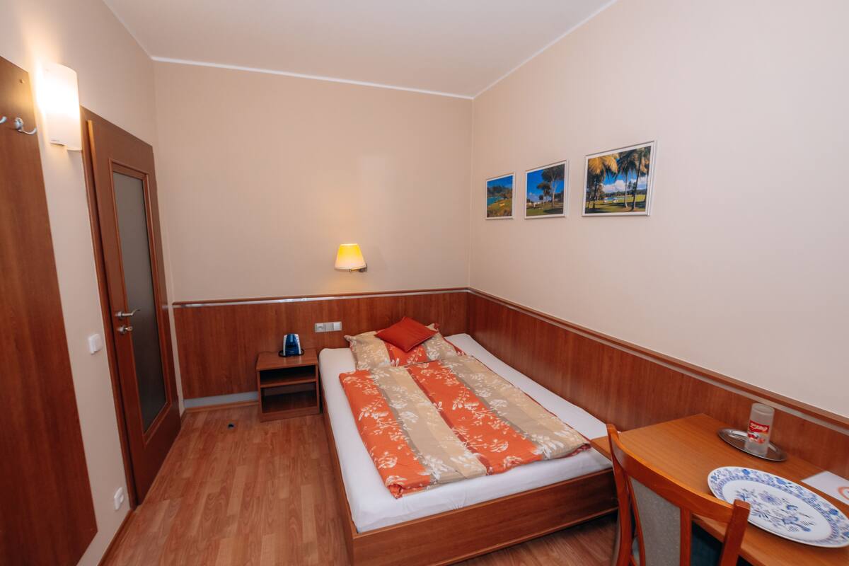 Quarto pequeno e bem iluminado, com cama de madeira, mesa de cabeceira e escrivaninha com cadeira. Três quadros emoldurados decoram a parede.