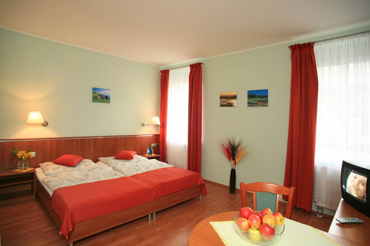 Quarto de hotel com duas camas, uma mesa de jantar com frutas, televisão e janela com cortinas.