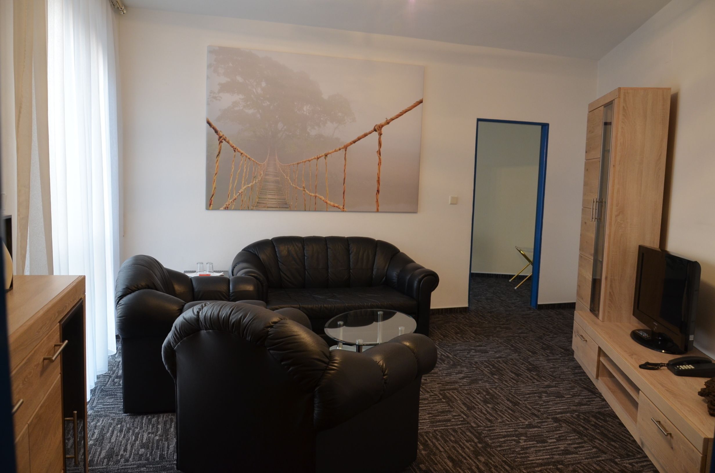 A modern living room with a black leather sofa, a glass coffee table, and a wooden cabinet with a television. A large abstract painting is on the wall, and there is a door leading to another room.
