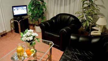 A living room with a black leather sofa, a glass coffee table with a tray of fruit and juice, a potted plant, a television on a stand, and a lamp on a side table.