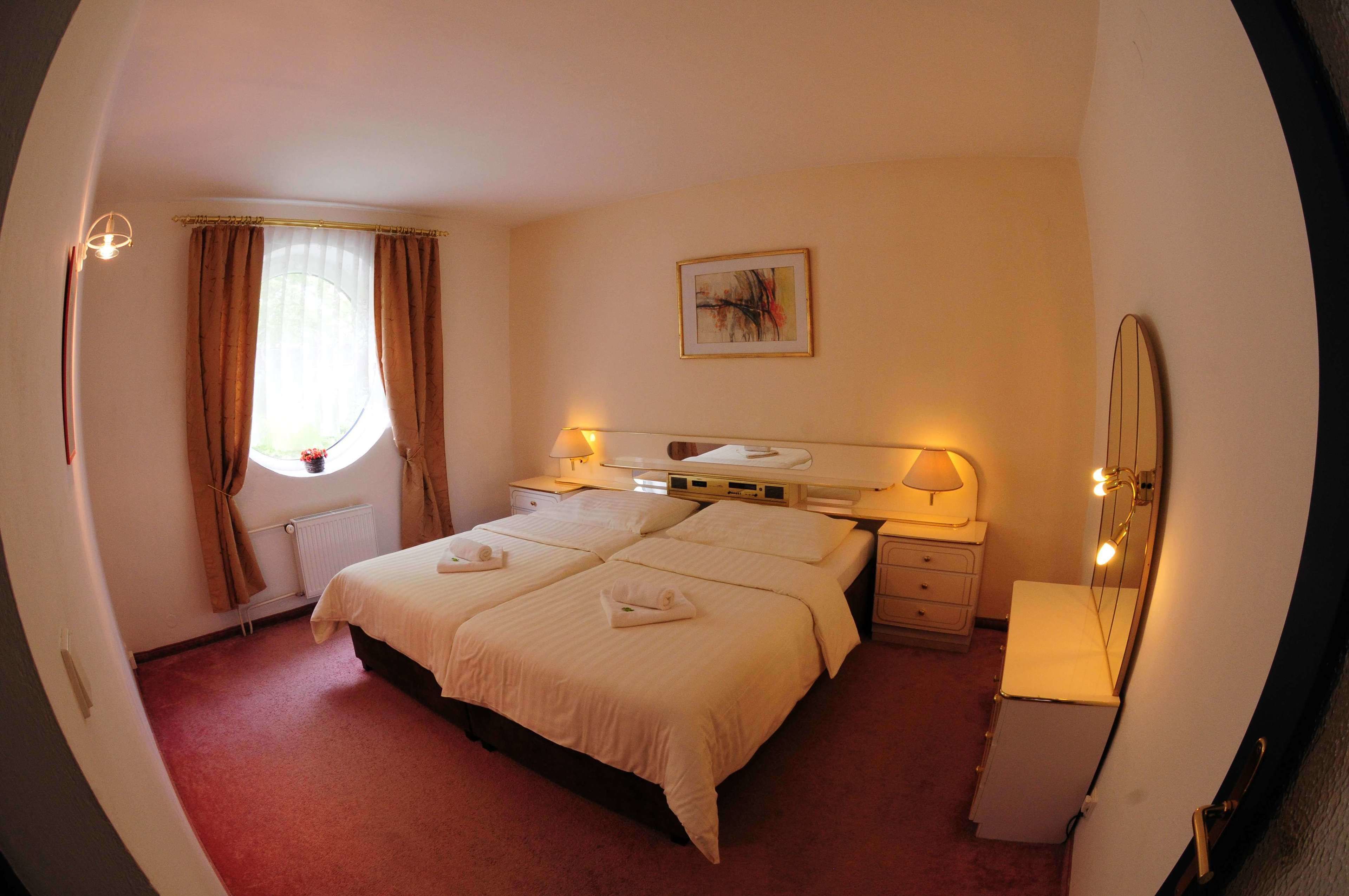 A hotel room with a large bed, bedside lamps, a window with curtains, a radiator, and a painting on the wall.