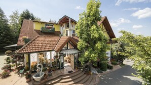 Un bâtiment à deux étages, doté d’un toit en tuiles, d’une entrée couverte et d’un jardin agrémenté de plantes en pot et d’une fontaine.