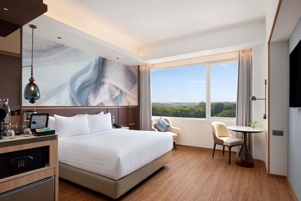 A hotel room with a bed, a desk with a coffee machine, a chair, a small table, and a large window with a view.