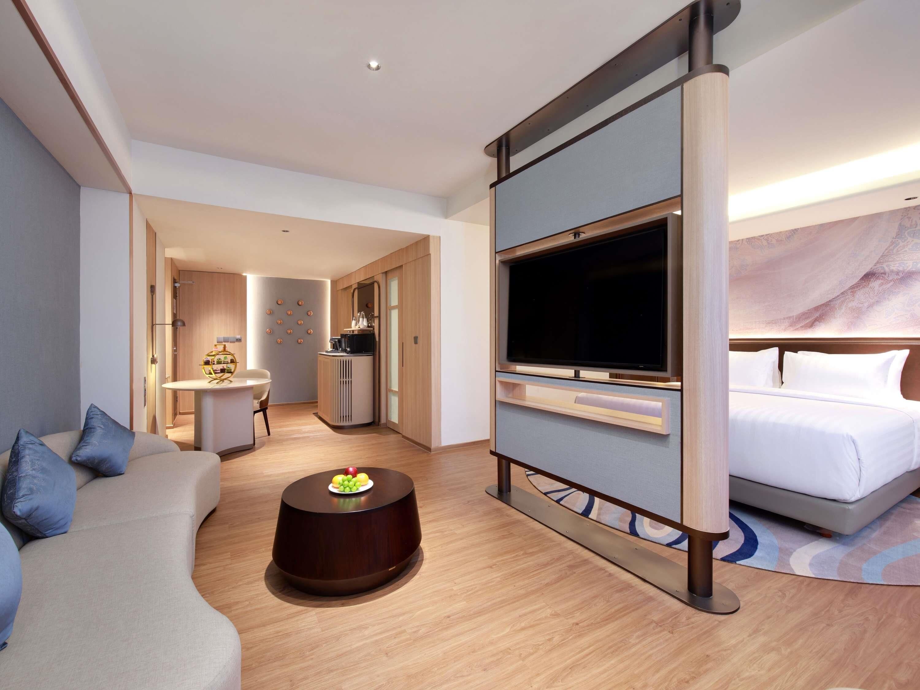 A modern hotel room with a sofa, a TV, a bed, a small dining area, and a bathroom.