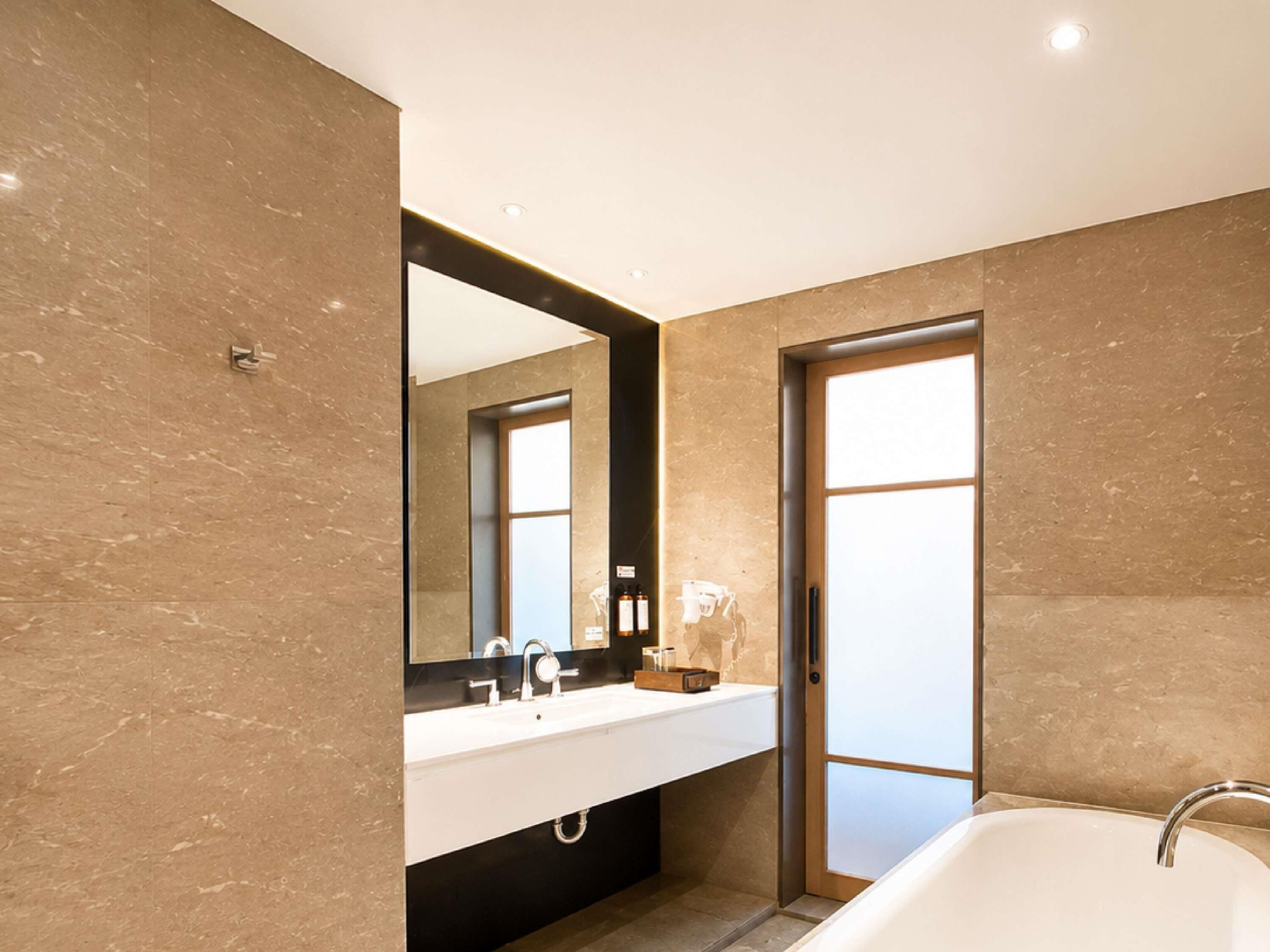 Un bagno moderno con uno specchio grande, un lavandino bianco, una vasca da bagno e una porta che dà su un'altra stanza.