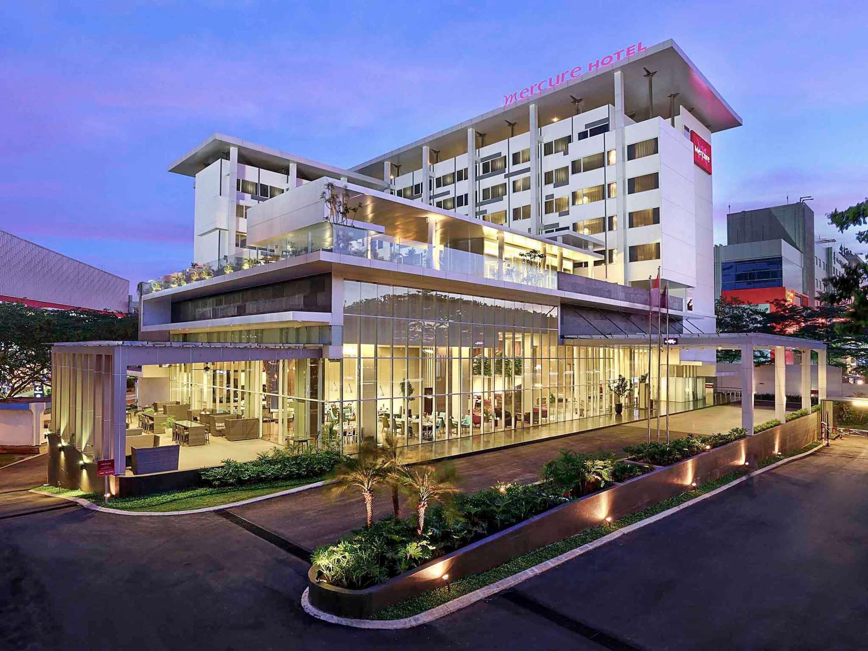 A modern hotel with a glass facade, outdoor seating area, and a visible name sign.