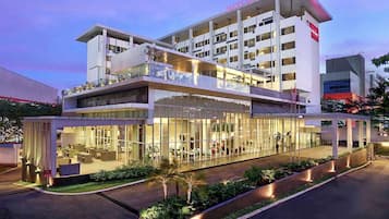 A modern hotel with a glass facade, outdoor seating area, and a visible name sign.