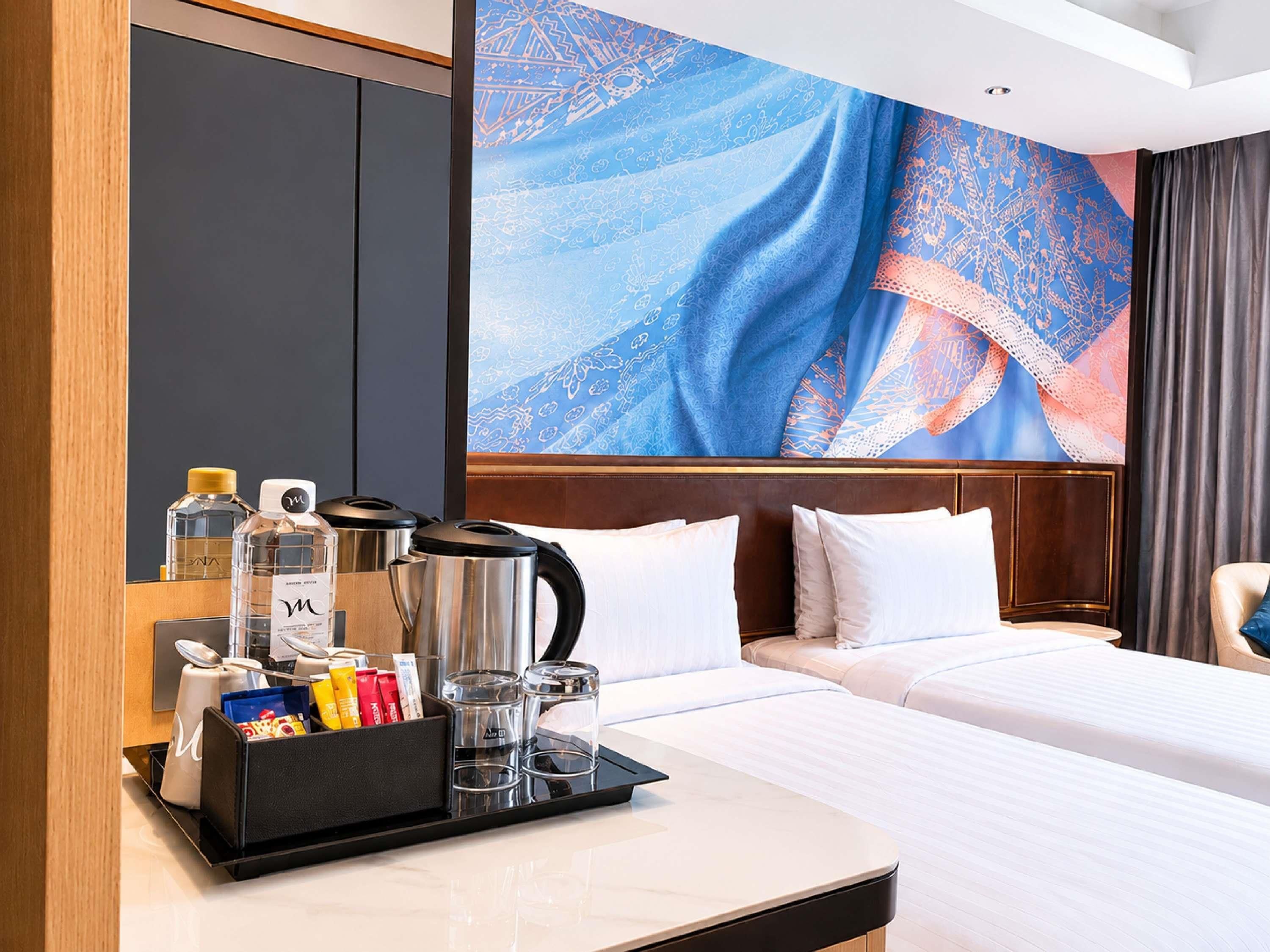 A hotel room with a bed, a bedside table with a coffee maker, a tray with toiletries, and a large abstract wall mural.