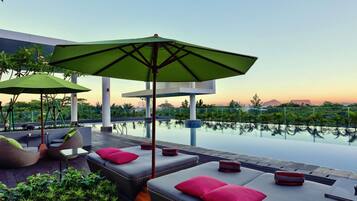 A rooftop pool area with lounge chairs and umbrellas, surrounded by greenery and overlooking a cityscape.