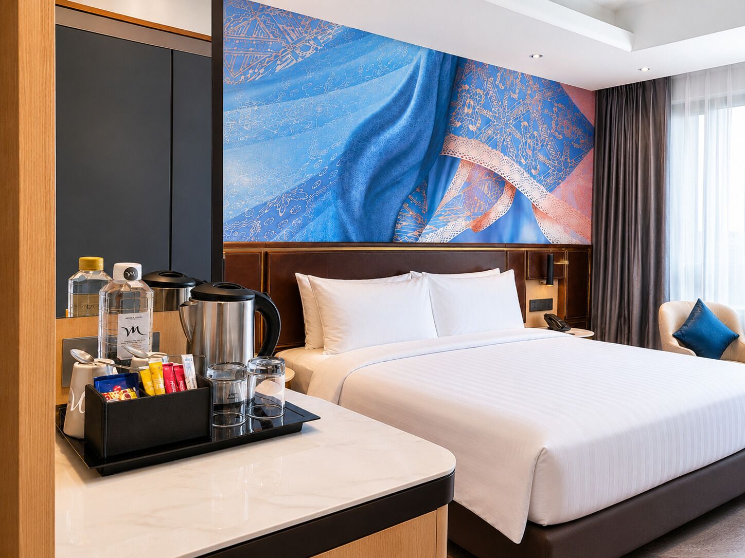 A hotel room with a bed, a bedside table with a tray of toiletries, a large patterned wall, a window with curtains, and a chair with a cushion.