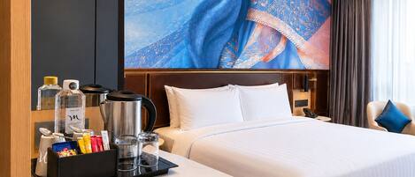 Une chambre d’hôtel avec un lit, une table de chevet sur laquelle se trouve une petite table avec des articles de toilette, un grand mur orné d’un motif, une fenêtre avec des rideaux et une chaise avec un coussin.