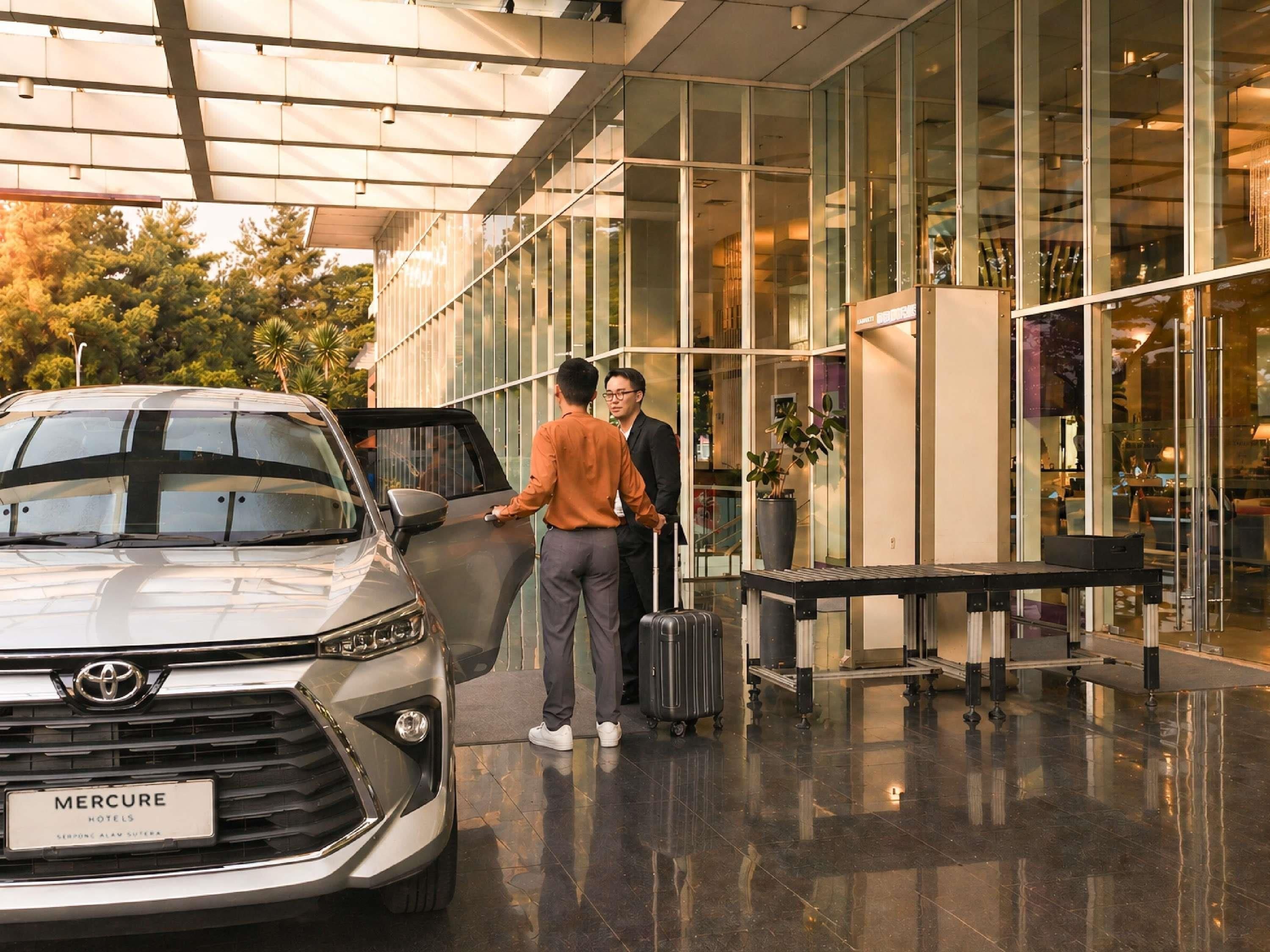 Entrada moderna de hotel com um carro estacionado em frente, um carrinho de bagagem e fachada de vidro.