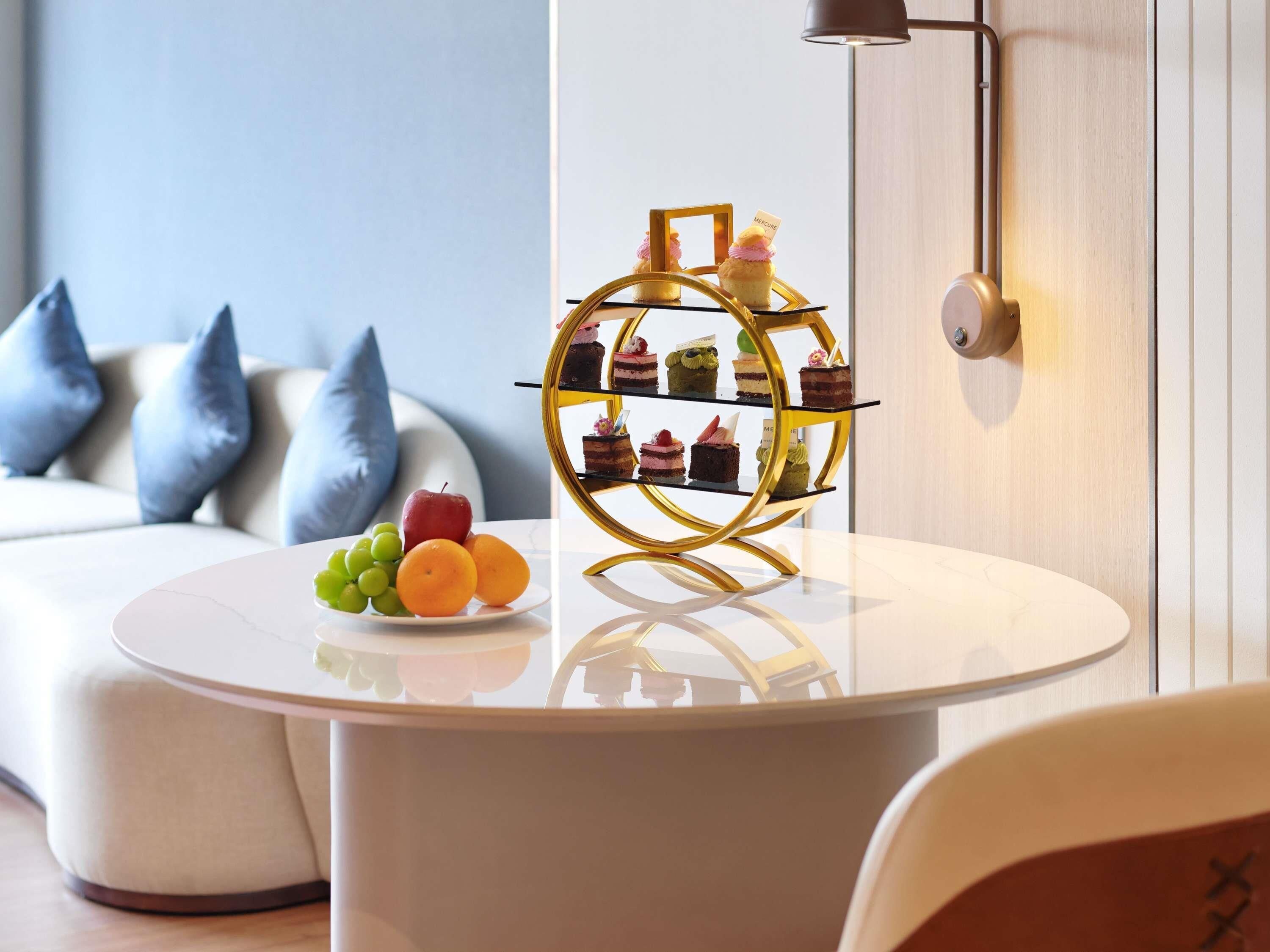 A round table with a fruit plate, a gold dessert stand, and a modern sofa with blue cushions.