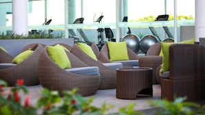 Un área de estar al aire libre de diseño moderno con mobiliario de mimbre y cojines verdes, con vista a un gimnasio con equipo de ejercicios.