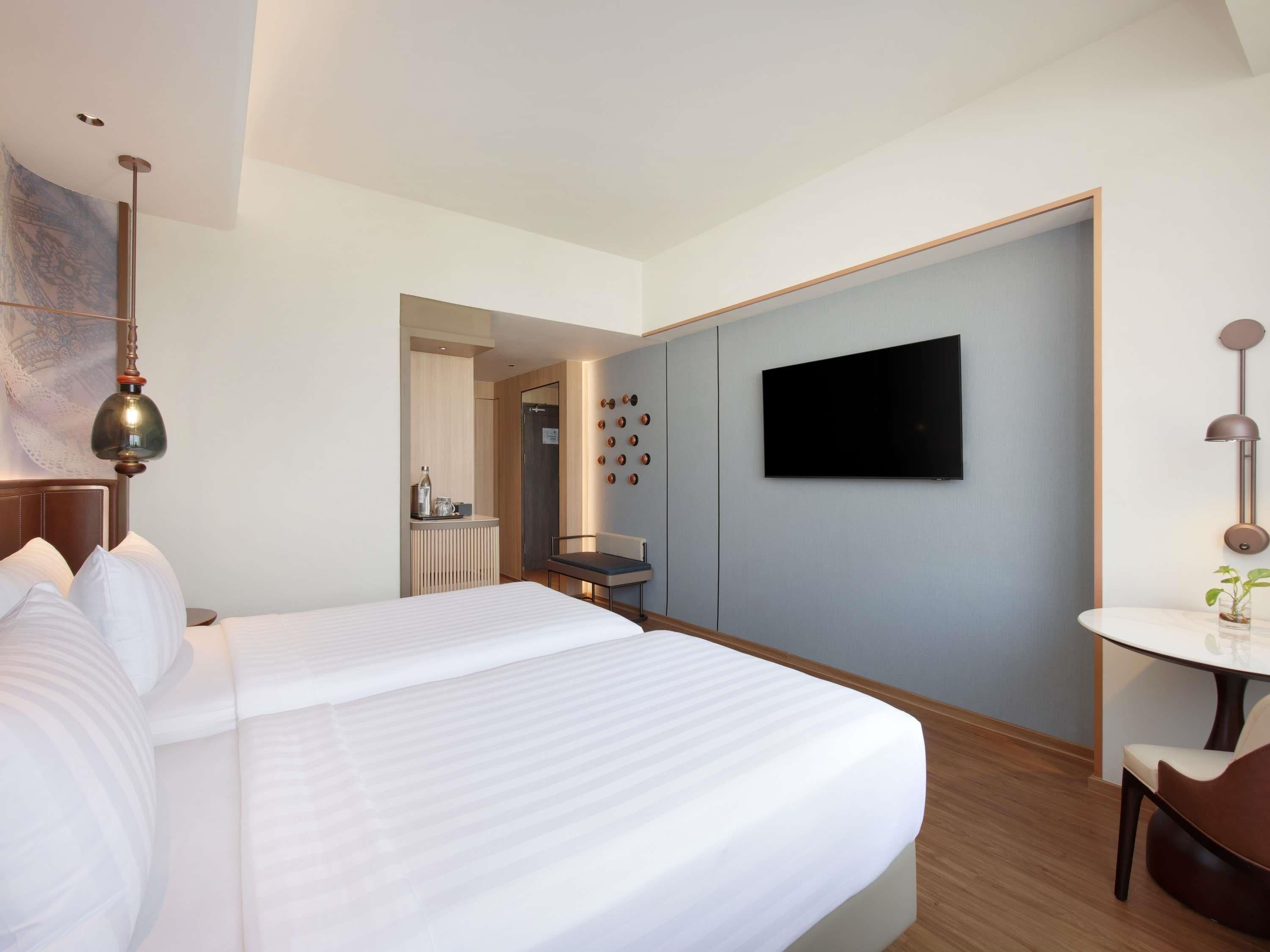 Habitación de hotel con una cama grande, un televisor montado en la pared, un escritorio con una silla y una mesita con una planta.