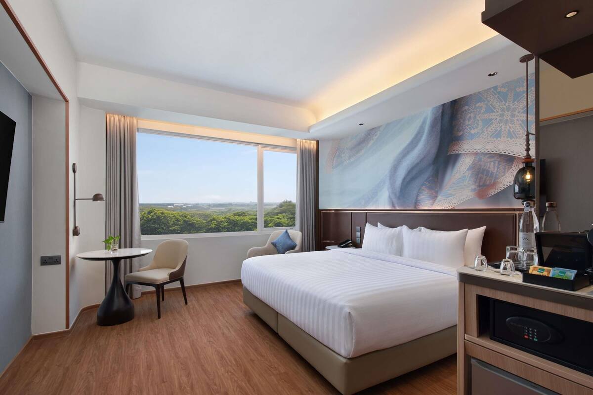 Habitación de hotel con cama, escritorio, silla, ventana con vistas y un cuadro en la pared.
