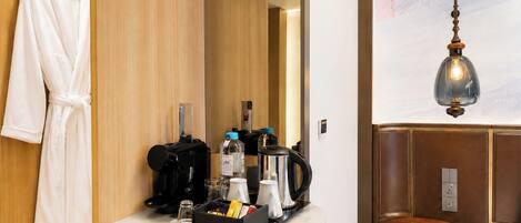 A hotel room with a robe hanging, a coffee machine, a bottle of water, a kettle, and a small table with a lamp.