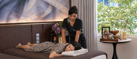 Ein Behandlungsraum mit Massageliege, einem Masseur und einem kleinen Tisch mit Blumenvase und einer Serviertschale mit Snacks.