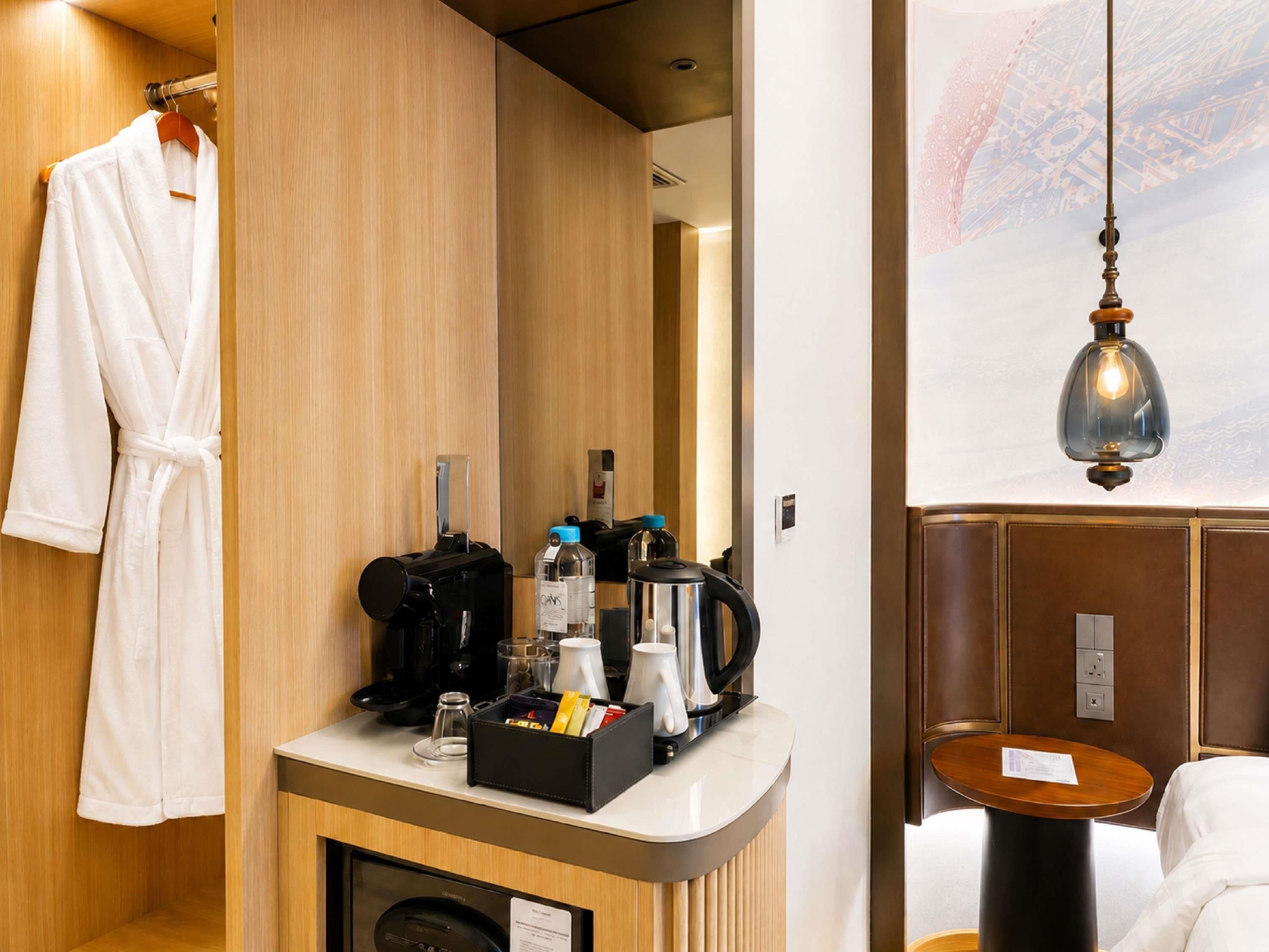 A hotel room with a wooden vanity, a robe hanging on the wall, a coffee maker, and a table with a lamp.
