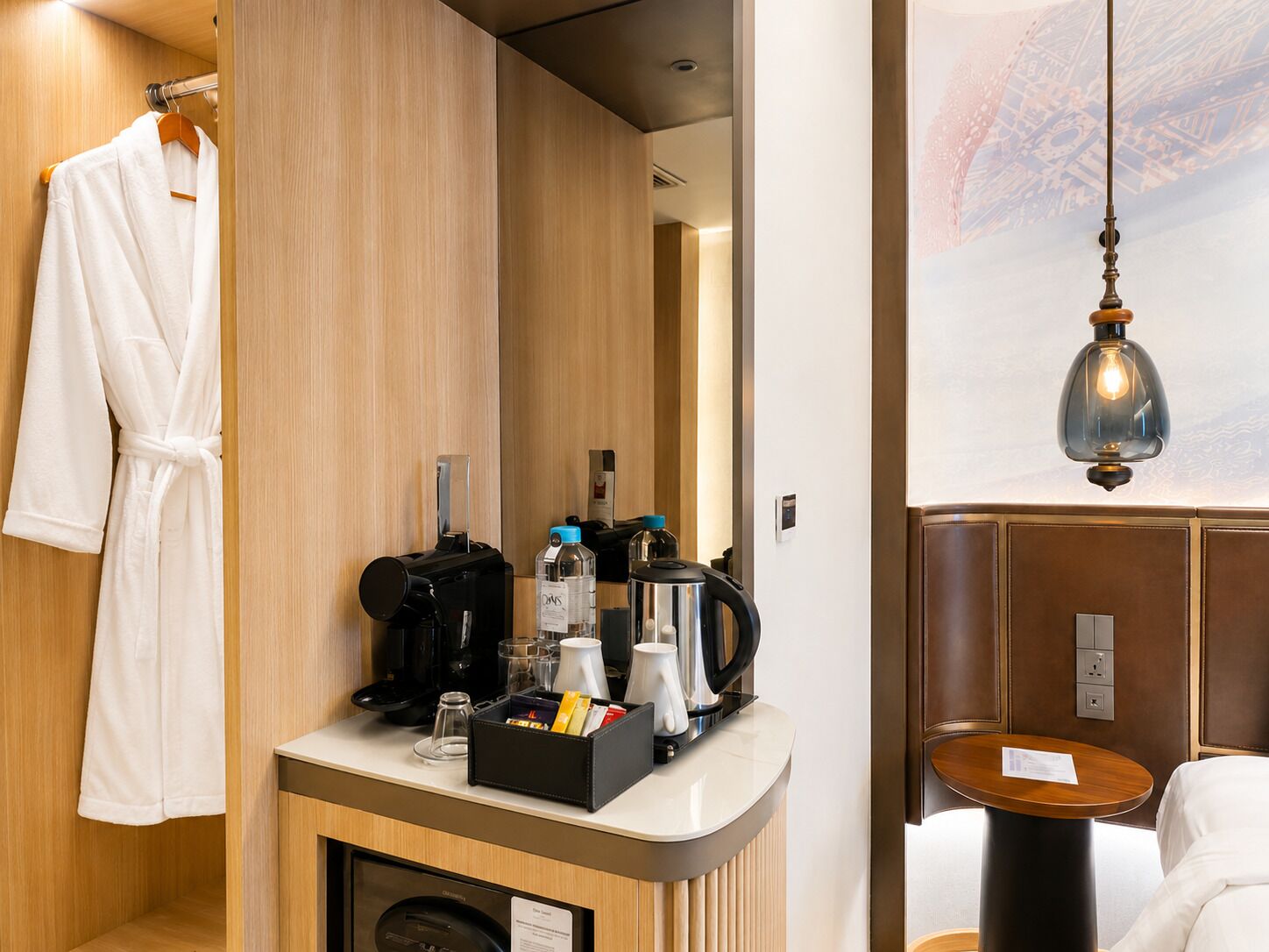 A hotel room with a wooden vanity, a robe hanging, a coffee machine, and a table with a lamp.